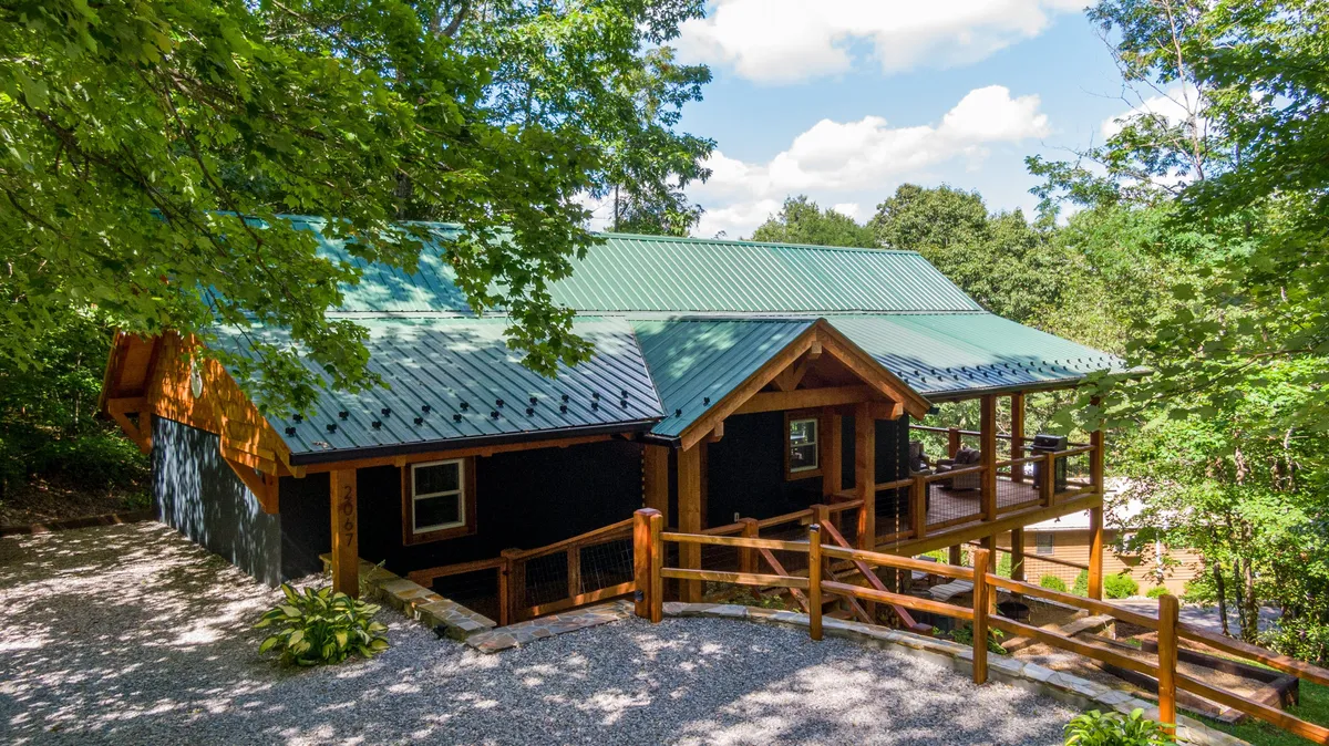The image showcases a charming single-family cottage nestled among mature trees, with a welcoming wooden porch, a striking green metal roof, and a spacious gravel driveway. The porch invites relaxation, while the surrounding woodland hints at adventure and privacy, all set in a tranquil, resort-style neighborhood in the Appalachian mountains.
