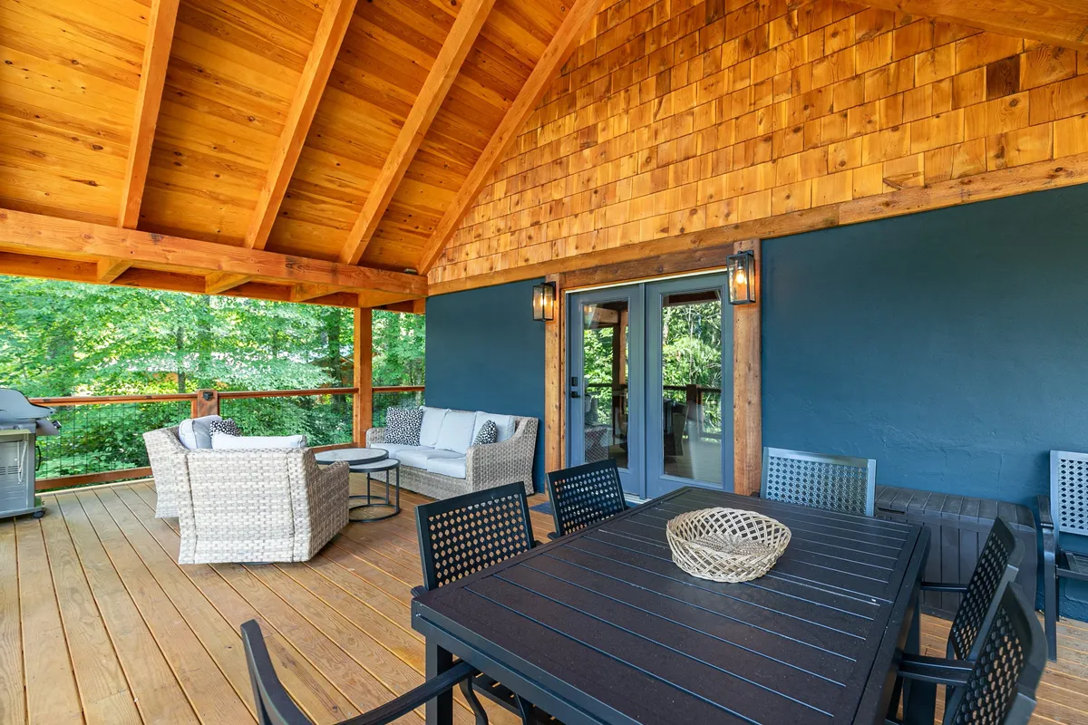 A serene outdoor dining area on a covered porch, featuring a large table for group meals, comfortable seating, a grill nearby, and rustic wooden beams. The space seamlessly connects with the natural surroundings, inviting guests to dine al fresco.