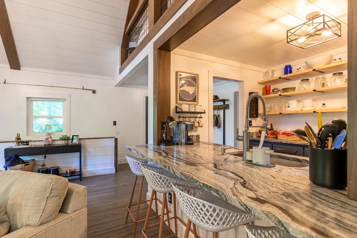 A bright, contemporary kitchen featuring a spacious island with bar stools, sleek shelving displaying dishware, and a stylish art piece on the wall. The layout is open and conducive to both cooking and socializing, with plenty of counter space and seating for guests or family members to gather around.