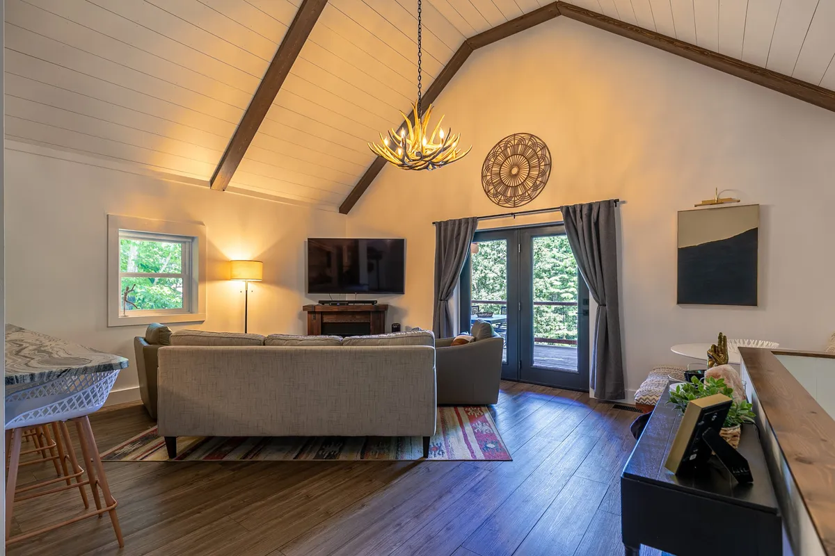 A cozy main living room with a plush sofa, stylish chandelier, large windows letting in natural light, a smart TV, and decorative wall art. The arrangement encourages relaxation and togetherness.