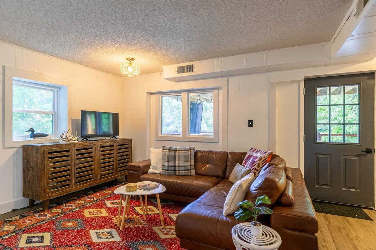 A spacious lower-level lounge with a large brown leather sectional, colorful area rug, wooden console table, TV, and windows bringing in daylight. The setup is ideal for casual gatherings, movie nights, or relaxing with friends and family.