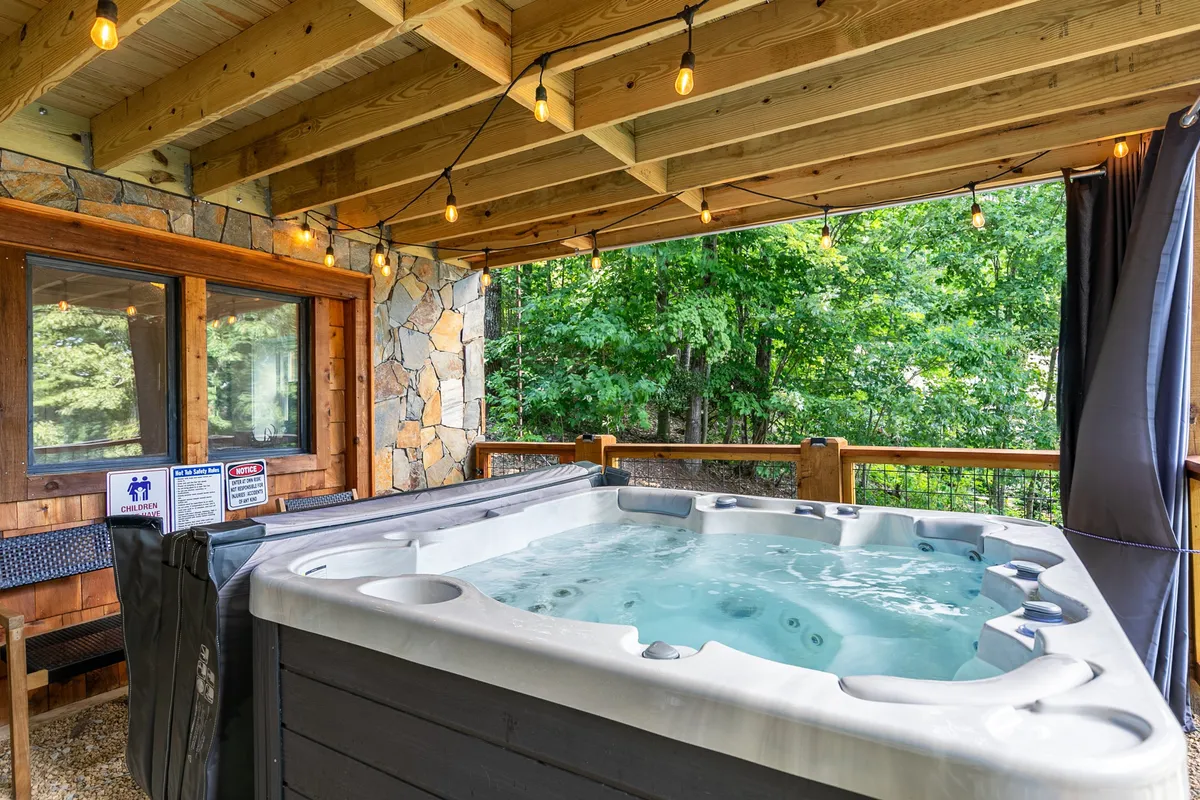 The lower patio area is shown at twilight, with a spacious hot tub, warm string lights, robust wooden beams, and a stone wall backdrop. The inviting setup is designed for both relaxation and social gatherings, emphasizing starlit soaks and evening ambiance.