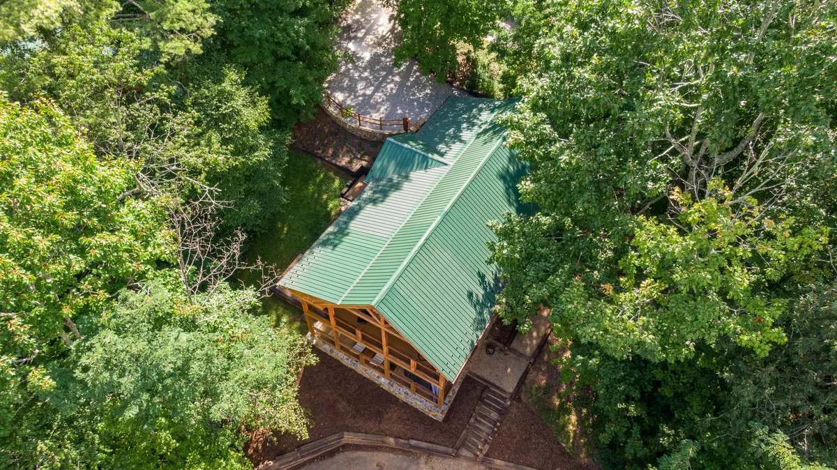 A scenic view of a cozy wooden cabin with a green metal roof, nestled among tall trees and fronted by a pathway. The porch offers a vantage point for sunrise views and stargazing.