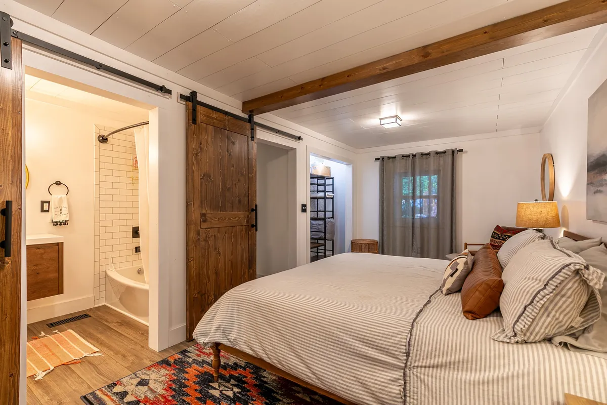 A serene bedroom featuring a cozy bed, sliding barn doors, a plush rug, and a window with soft curtains letting in natural light. The space feels relaxing and perfect for unwinding after a day in the mountains.