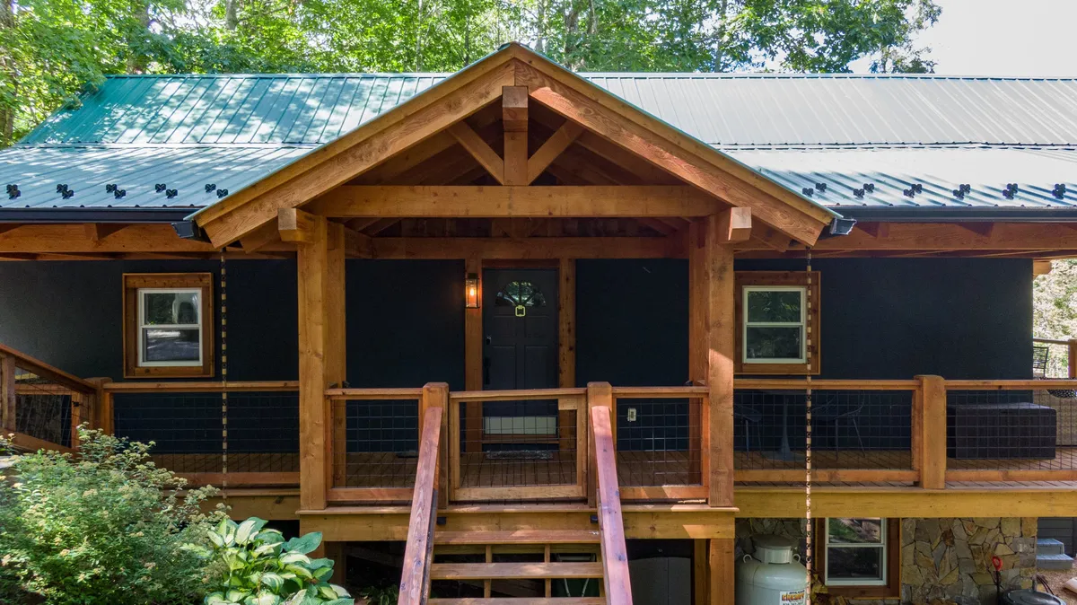 A welcoming wooden porch with a slanted roof, front door with decorative glass, and space for gathering. The area is set against a backdrop of natural greenery, hinting at mountain surroundings and offering a glimpse of the home’s connection to the environment.