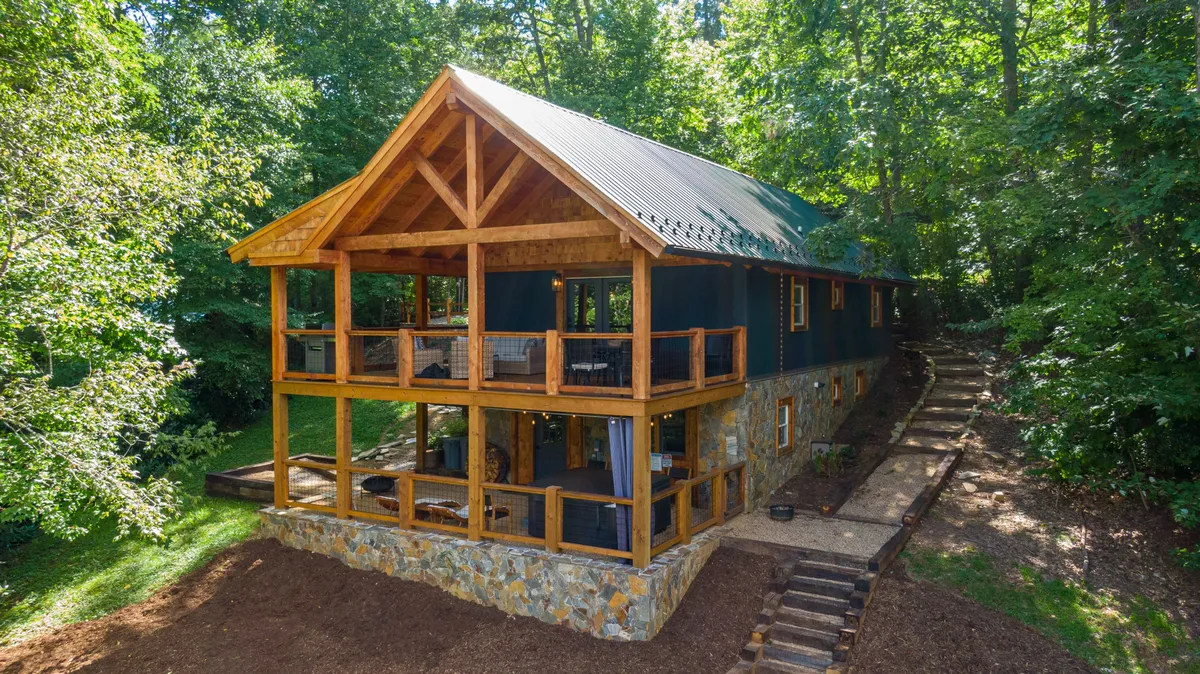 This image features a spacious wooden deck with a stone foundation, large windows, and surrounding trees. The deck offers ample room for outdoor gatherings and overlooks nearby nature trails, creating a seamless connection to the environment.