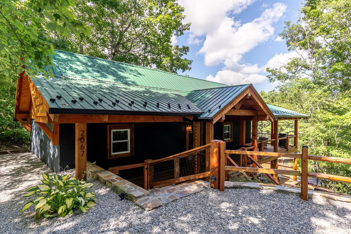 A wide view of the home’s expansive wooden porch wraps around the exterior, featuring a green metal roof, stone pathway, and lush greenery that frames the setting. The scene exudes the natural seclusion and peacefulness described in the section, emphasizing the porch as a central indoor-outdoor living space.