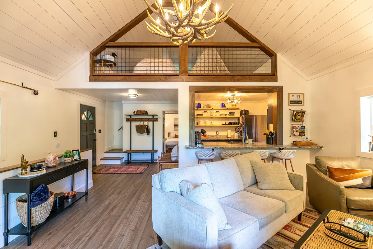 This image showcases an inviting open living space with a comfortable sofa, a bar counter with stools, and an antler chandelier. The open loft area above hints at the home's flexible sleeping arrangements, while decorative shelves and wood accents add warmth. The scene exudes a sense of relaxed gathering and is perfect for both coffee mornings and lively evenings.