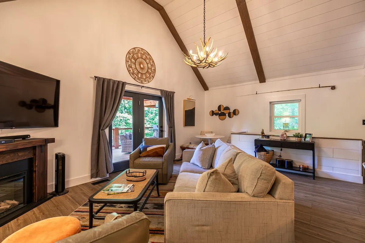 A spacious lounge area featuring a fireplace, large TV, ample sofa seating, and decorative chandelier. Large windows and an exterior door provide glimpses of the outdoors, reinforcing the connection between indoor relaxation and outdoor activities. The space is ideal for movie nights, board games, or relaxing after outdoor fun.